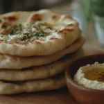 Stacked homemade flatbread with hummus in a rustic kitchen setting.